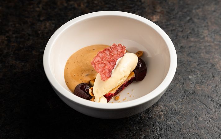 Dessert in weißer Schale mit Karamellcreme, Kirschen und rosa Knusperblatt