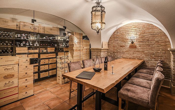 Gewölbekeller mit langem Holztisch, Polsterstühlen und Weinkisten hinter Glas